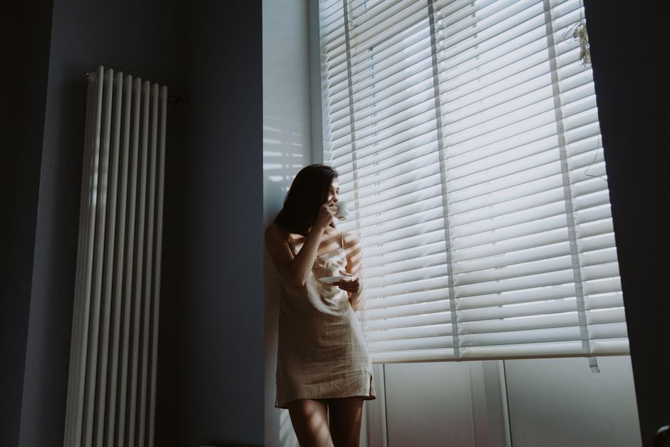 A woman in soft morning light stands by the window, holding a cup and saucer, savoring a peaceful moment.